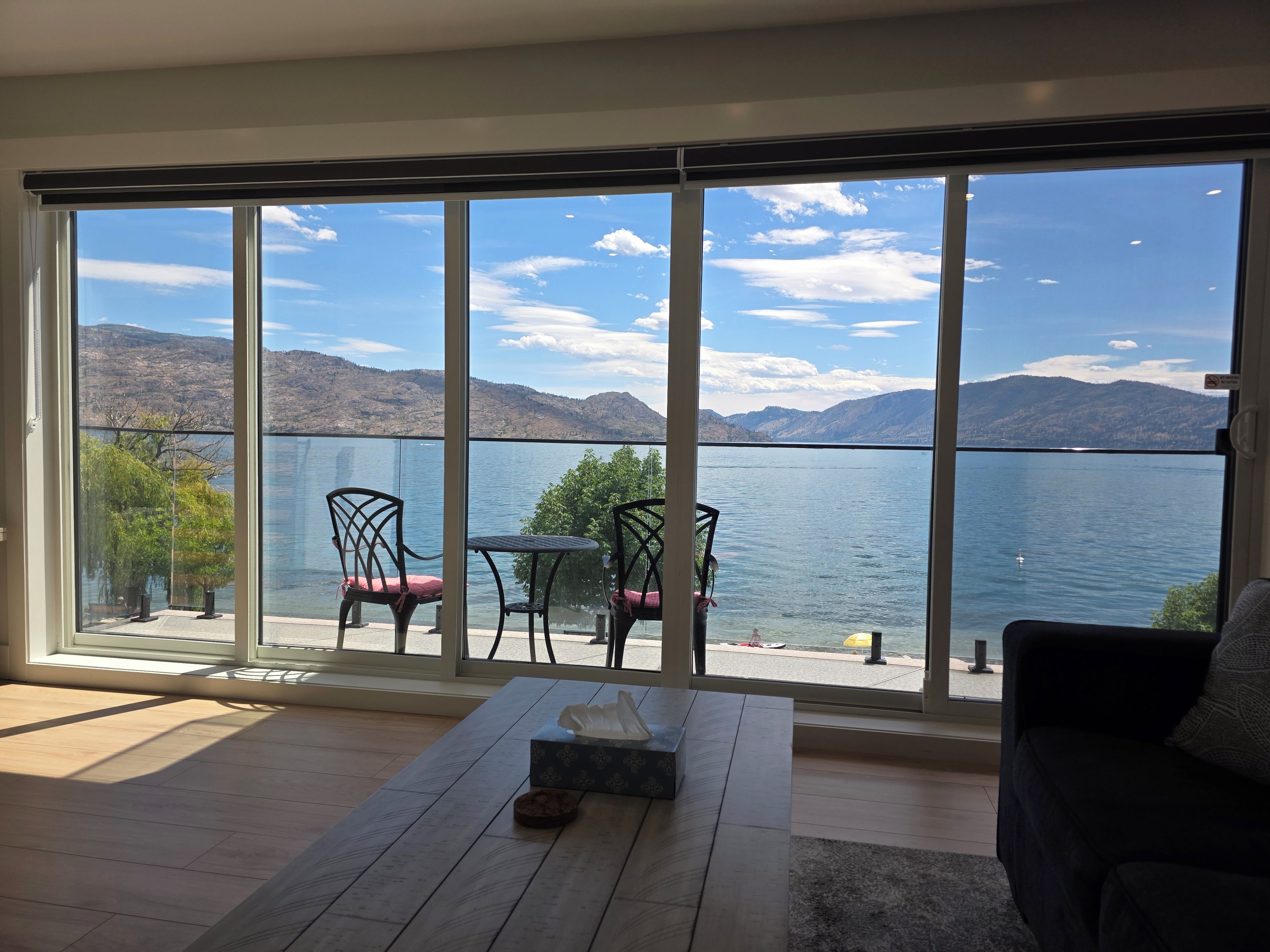 Una habitación con un gran ventanal corredero que da a una vista del lago. Adentro, se ve un suelo de madera, una mesita con una caja y un sofá de color oscuro.