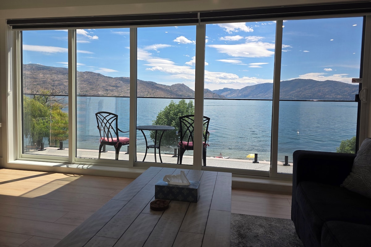 Quarto com uma grande porta de correr de vidro que dá para uma vista do lago. No interior, há piso de madeira, uma pequena mesa com uma caixa e um sofá de cor escura.