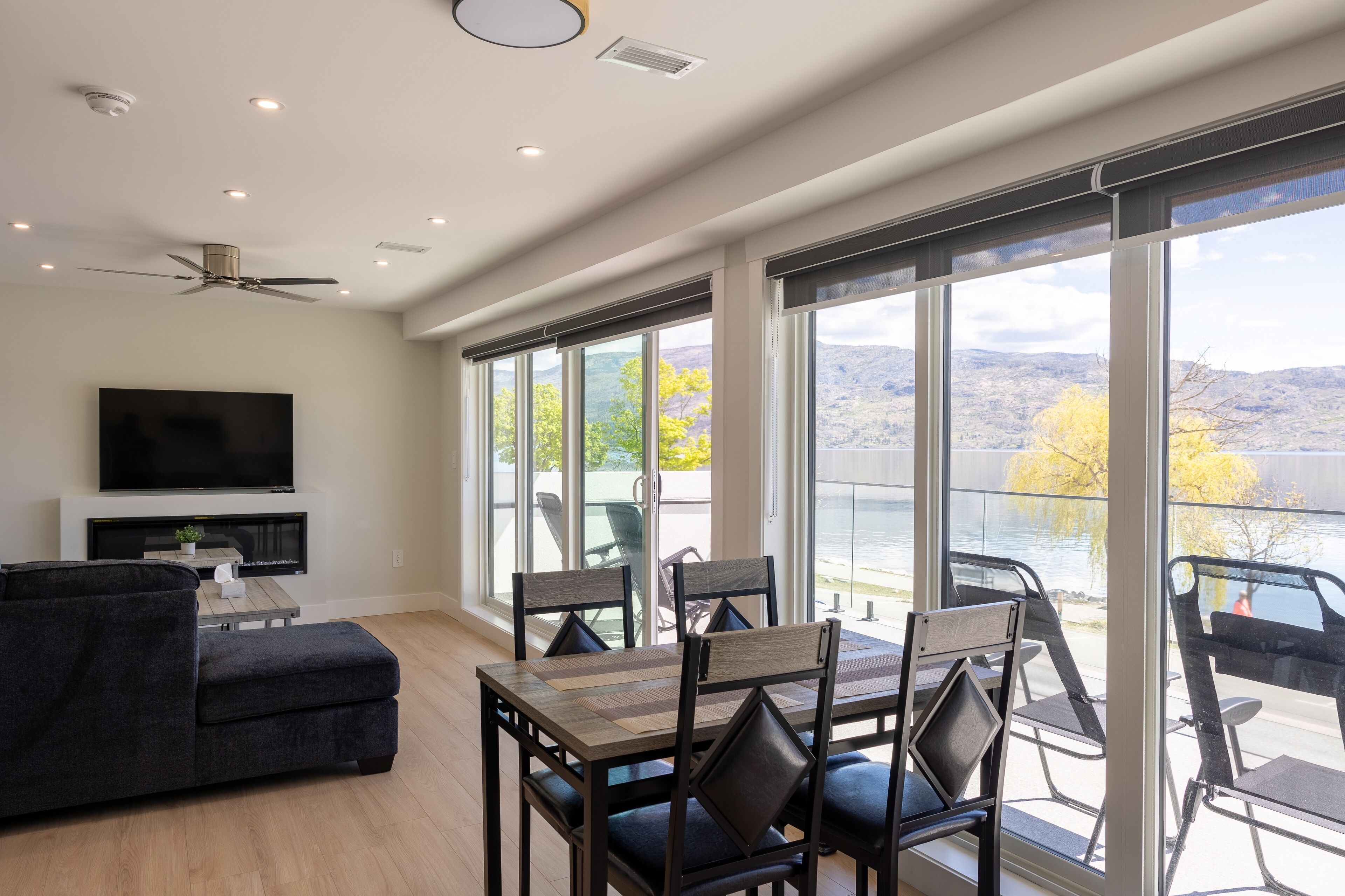 Una sala moderna con televisor de pantalla plana, sofá de color oscuro y un comedor con mesa y sillas. Ventanas amplias ofrecen vista a un lago y montañas.