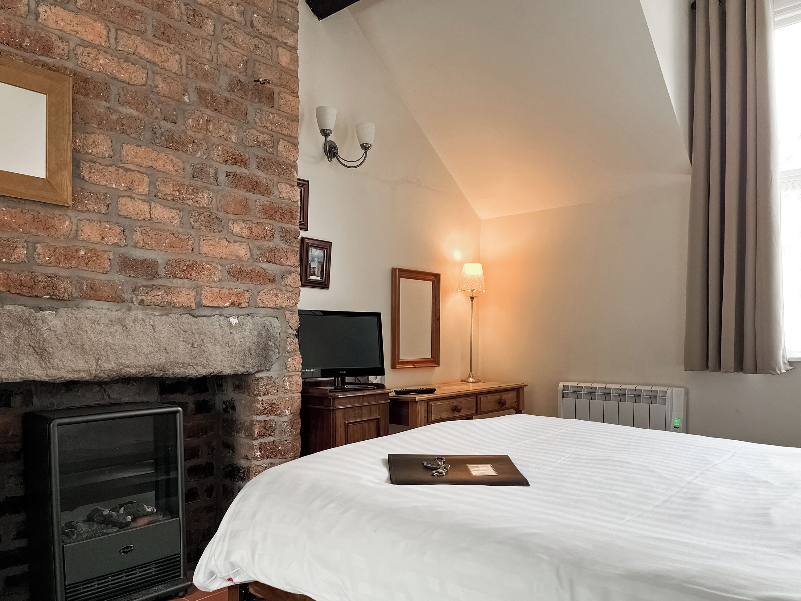 A bedroom with a brick fireplace, a flat-screen TV, a bedside table, and a bed with a white comforter.