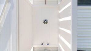 Une douche extérieure blanche, dotée d’un robinet en métal et de deux poignées bleues.