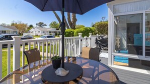 Une table à plateau de verre, sur laquelle sont posés un pot de fleurs et un coquillage, se trouve sous un grand parasol, sur une terrasse en bois dotée de rampes blanches, et offre une vue sur une maison et des voitures au loin.