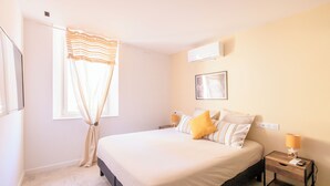 A bedroom with a bed, bedside lamps, a window with curtains, an air conditioner, and a wall-mounted picture. - Magnifique 6 Personnes Avec Terasse Tropezienne Refait à Neuf (Bandol)