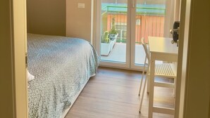 Ein Schlafzimmer mit Bett, einem kleinen Tisch und einer Schiebetür, die auf einen Balkon mit Blick auf ein Gebäude und einen Zaun führt.