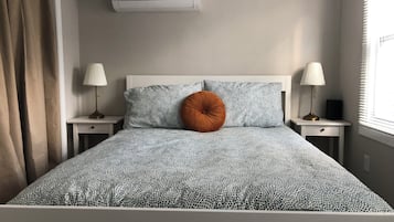 Cama bem arrumada com roupa de cama estampada, duas luminárias de cabeceira, ar condicionado fixado na parede e uma janela com persianas.