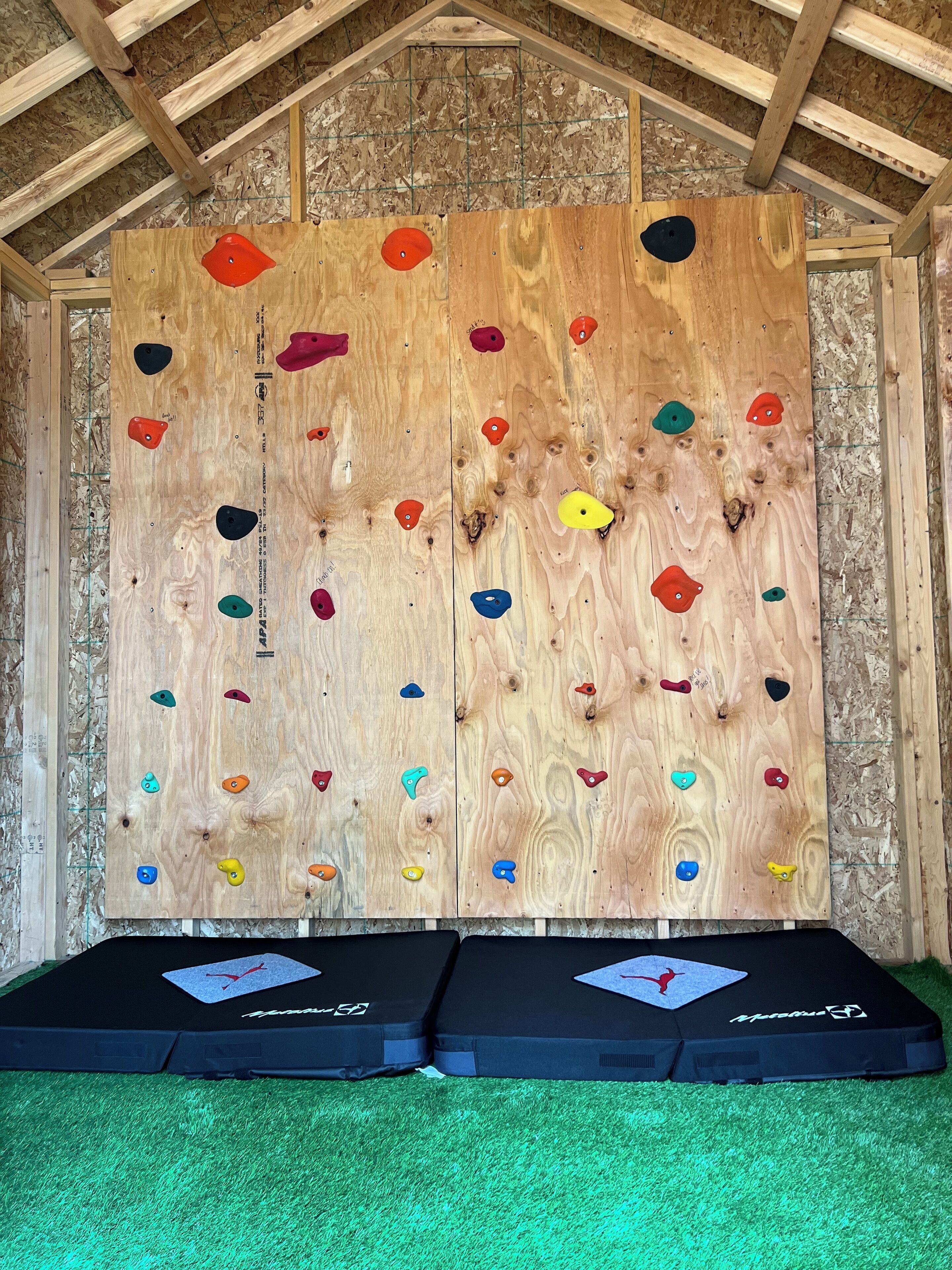 Una pared de escalada de madera con presas de distintos colores y dos colchonetas negras en la base.
