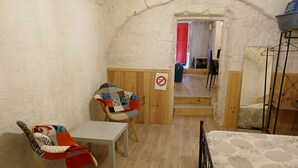 A small, cozy room with a wooden door, a white table, two chairs with colorful cushions, a bed with a patterned bedspread, and a small shelf with a basket. - Studio Dans Voûtes (Creissels)