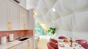 Een moderne keuken met witte kastjes en gele handgrepen, een eethoek met roze stoelen en een tafel gedekt voor vier personen, en een grote tentachtige constructie op de achtergrond.