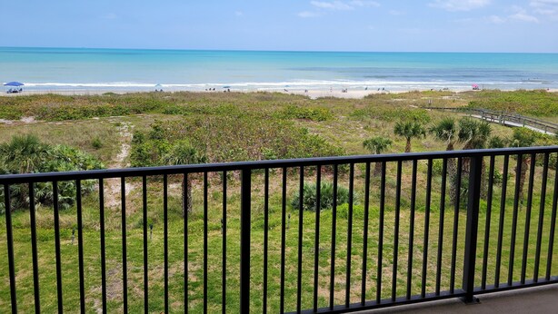 A balcony with a view of a beach, a wooden walkway, and some greenery. - Ocean Front Condo Downtown Cocoa Beach (Cocoa Beach)