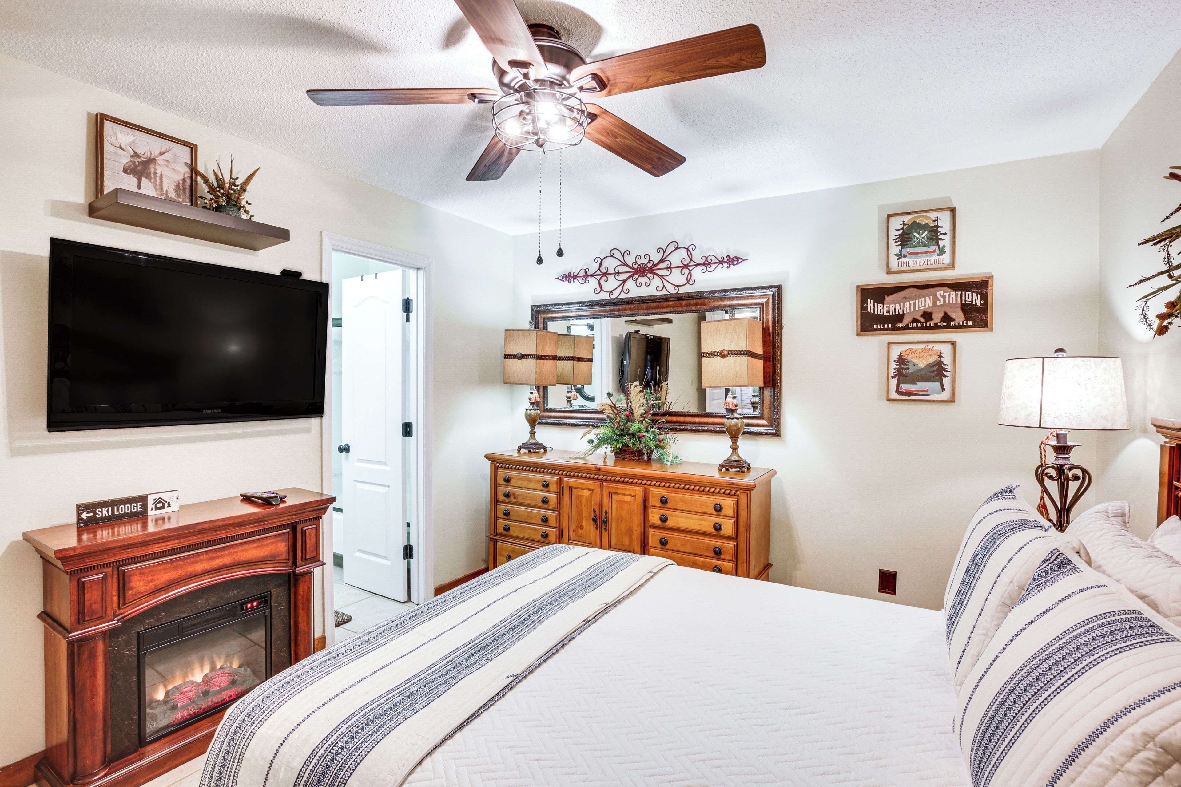 A bedroom with a bed, a fireplace, a television, a ceiling fan, a dresser, a mirror, and framed pictures on the wall.
