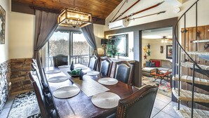 Un comedor con mesa de madera, sillas y una araña. Se observa una escalera con barandilla de fibra y vistas al exterior.