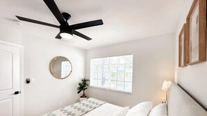 Ein Schlafzimmer mit einem Bett, einem Deckenventilator, einem Fenster mit Jalousien, einem runden Spiegel und einem Nachttisch mit Lampe.