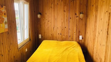 Ein Zimmer mit Holzvertäfelung, einem Bett mit gelber Bettdecke, einem Fenster und zwei Wandleuchten.