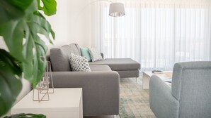 Una sala moderna con un sofá gris, una mesa de centro y una mesita auxiliar. Hay una planta en primer plano y una lámpara colgando del techo.