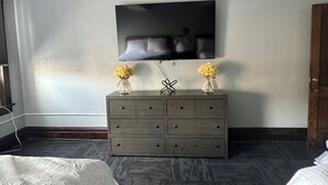 Una camera d'albergo con una grande TV a schermo piatto montata a parete, un comò in legno con due vasi di fiori e un letto con copripiumino bianco.