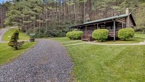 A rustic log cabin with a porch, surrounded by a gravel path and lush greenery.