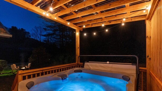 A hot tub under a wooden pergola with string lights, surrounded by a wooden deck.