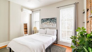 A bedroom with a bed, a ceiling fan, a window with curtains, a wall-mounted air conditioner, a painting, and a potted plant. - In the heart of Adams Morgan | Vintage Charm (Washington)