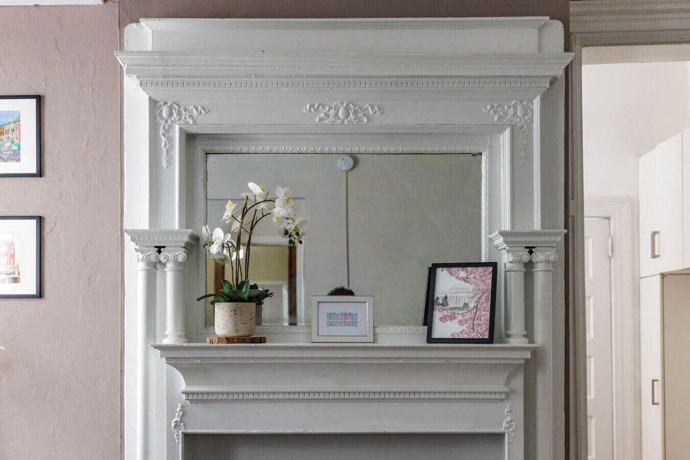 Une cheminée blanche surmontée d’un miroir, flanquée de deux colonnes. Une image encadrée est posée sur le manteau de la cheminée, et un pot de fleurs avec des fleurs blanches se trouve à proximité.