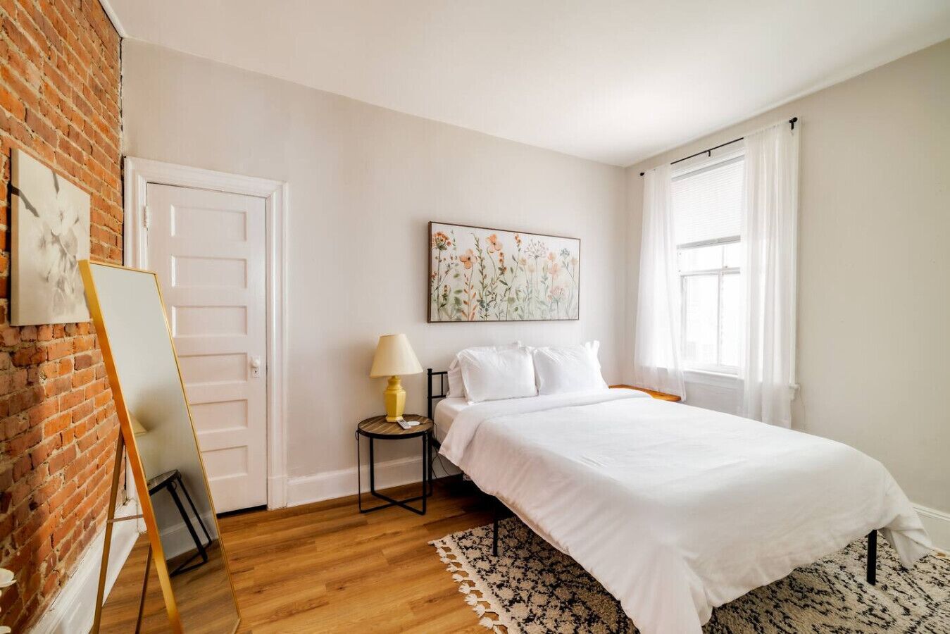 A bedroom with a wooden floor, a bed with white bedding, a small bedside table with a lamp, a framed picture on the wall, and a window with curtains.