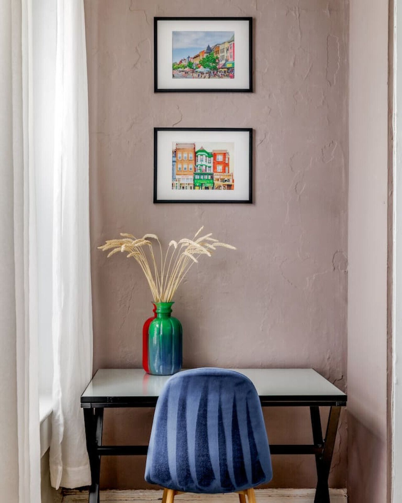 Una habitación con un escritorio, una silla, un florero con plantas secas y dos cuadros en la pared.