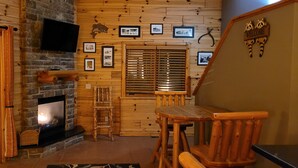 A cozy cabin interior with a fireplace, wooden walls, a dining area, and a 'Welcome' sign.
