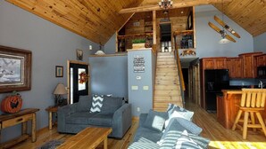 A spacious living area with a high wooden ceiling, a grey sofa, a wooden coffee table, and a kitchen area with wooden cabinets and a bar.