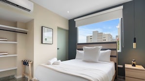 Ein modernes Hotelzimmer mit Bett, Nachttisch, Kleiderschrank und Fenster mit Blick auf die Stadt.