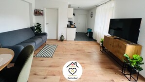 Un salón moderno con un sofá gris, suelo de madera, un televisor sobre un mueble de madera y una pequeña mesa con sillas.