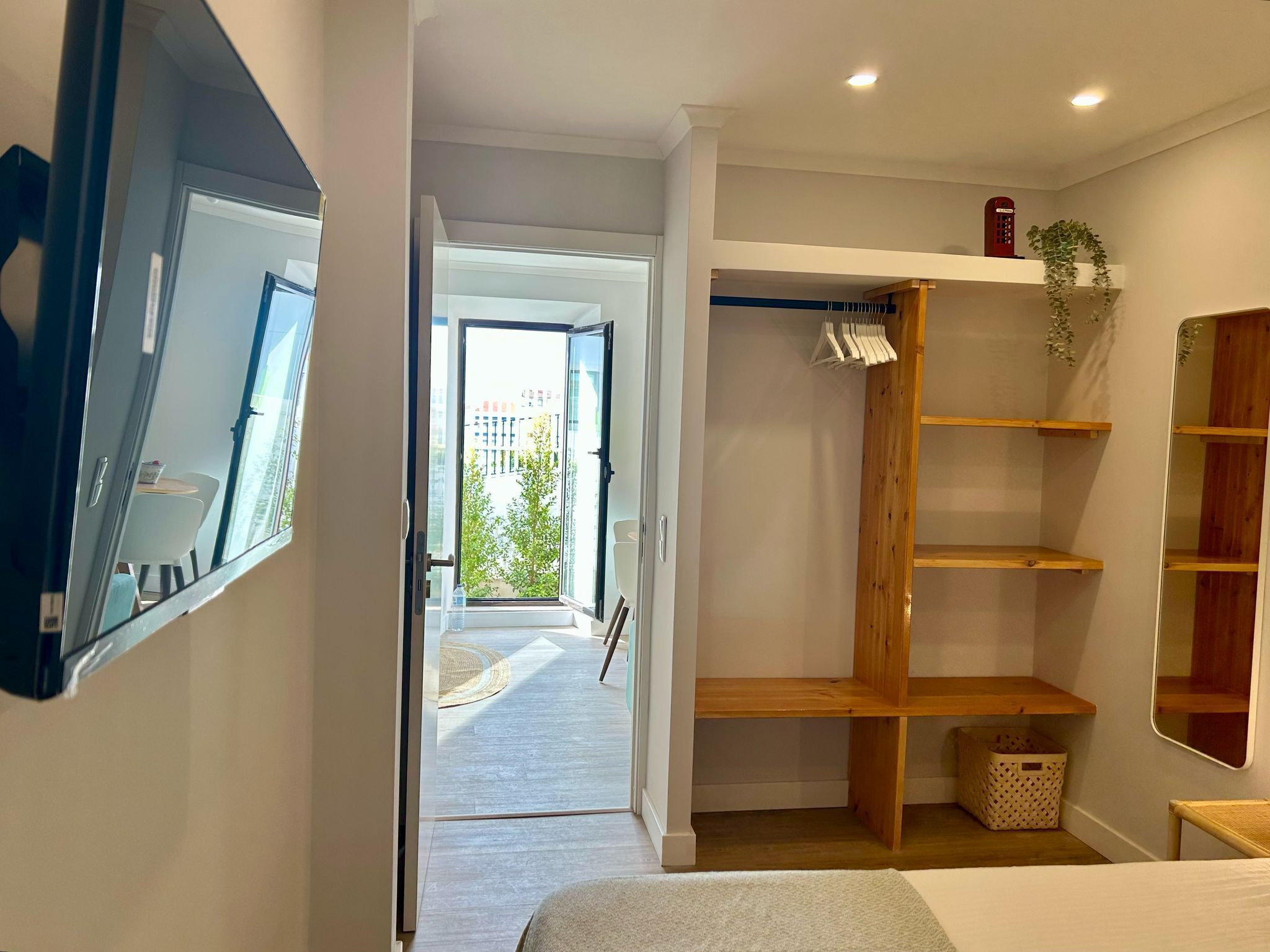 A modern bedroom with a large mirror, a wardrobe with shelves, a small table, and a view of the outdoors.
