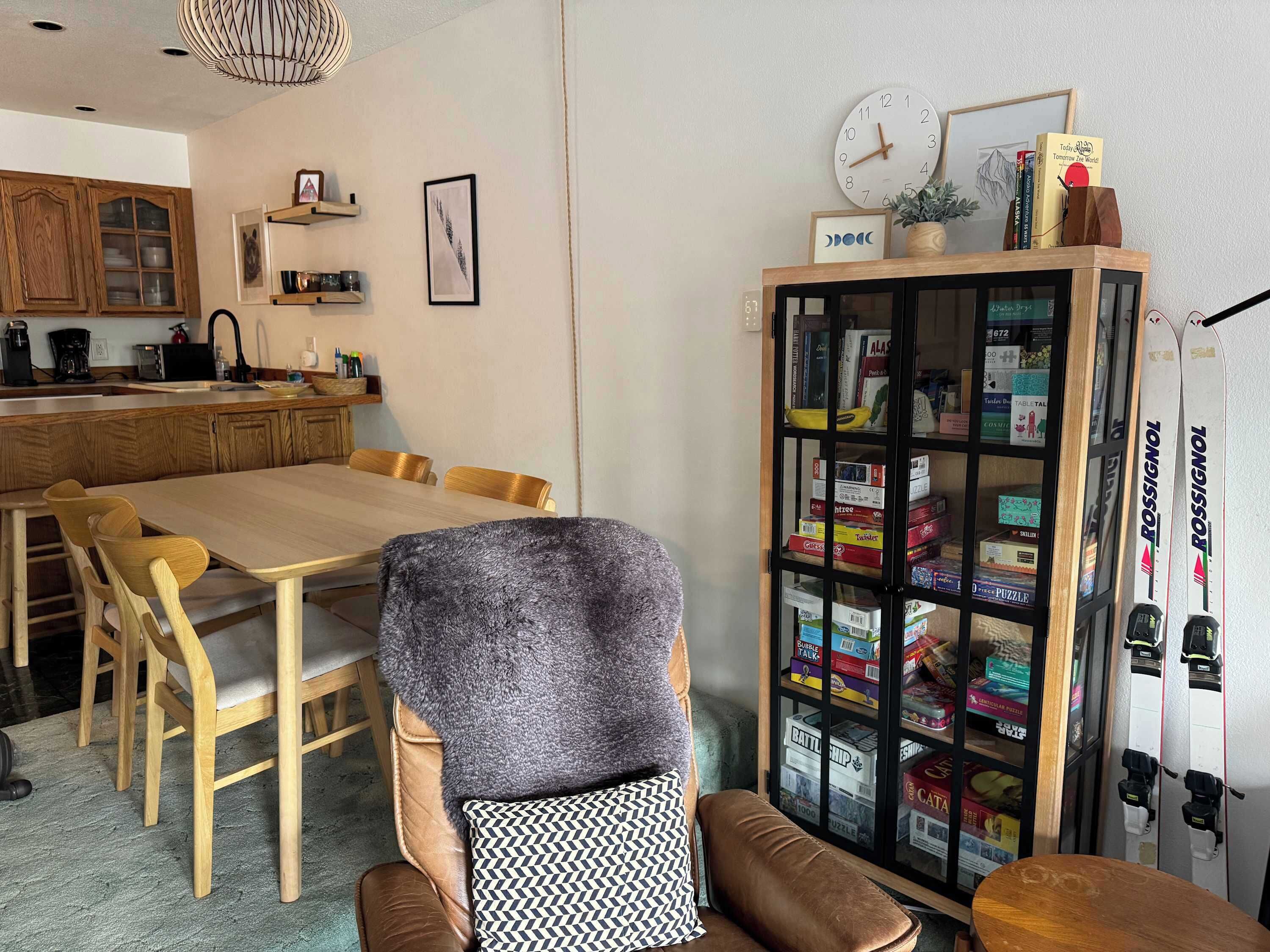 A cozy dining area with a wooden table and chairs, a bookshelf with puzzles, and a clock on the wall.