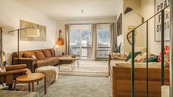 A cozy living room with a brown sofa, a wooden coffee table, and a stone fireplace. There is a staircase with a railing leading to a balcony.