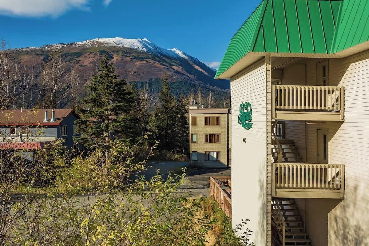 A building with a green roof and a balcony, surrounded by trees and a mountain in the background.