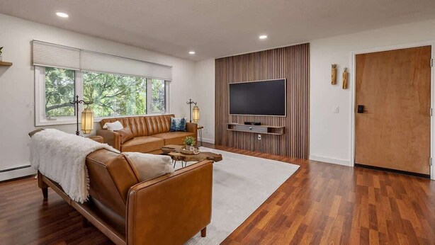 Una sala de estar moderna con un sofá de cuero marrón, un televisor de pantalla plana montado en una pared de madera y un ventanal con persianas.