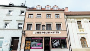 Un bâtiment sur lequel on peut lire l’inscription « UHREN-BURGMAUER », avec des devantures de magasins portant les enseignes « VAPE SHOP » et « DAMPFBEHANDLUNG VOR DRY ».