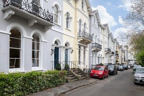 A row of traditional townhouses with white facades, black iron balconies, and a narrow street lined with parked cars.