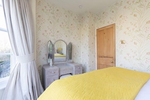 Un dormitorio con papel tapiz floral, una puerta de madera, un tocador con espejo y una colcha amarilla.