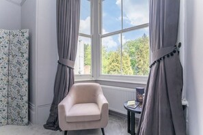 A room with a large window, a chair, a radiator, and a small table with a book and a candle.