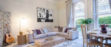 A living room with a sectional sofa, a coffee table, a dining table with chairs, a chandelier, and a large window with curtains.