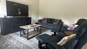 A living room with a dark wooden coffee table, a black sofa with patterned cushions, and a flat-screen TV on a black cabinet.
