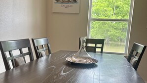 A dining table with a clear glass pitcher and a woven coaster, surrounded by four chairs. A framed picture of a boat is on the wall.