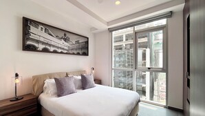 Un dormitorio moderno con una cama grande, un ventilador de techo, una ventana con vistas a un edificio y un cuadro en la pared.