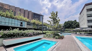 Un área de piscina en la azotea con dos piscinas, rodeada de cubiertas de madera y abundante vegetación.