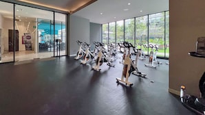 Un gimnasio con bicicletas estáticas y equipamiento cardiovascular, ventanas amplias y una entrada de cristal.