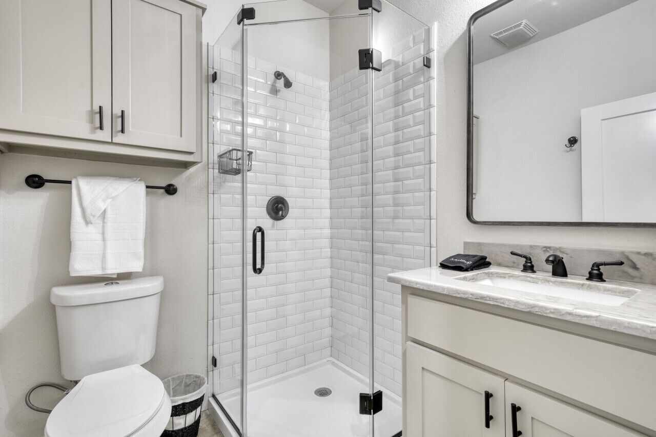 Un baño moderno con ducha envuelta en vidrio, paredes cubiertas de azulejos blancos, un inodoro y un lavamanos con encimera de mármol.