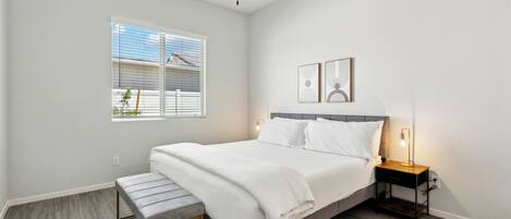 A bedroom with a bed, bedside table, bench, ceiling fan, window, and wall-mounted artwork.
