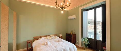 A bedroom with a bed, a chandelier, a dresser, a potted plant, and a window with a view.