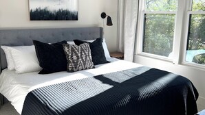 Ein Schlafzimmer mit Bett, Nachttisch, Wandlampe, einem Fenster mit Vorhängen und einem Bild an der Wand.