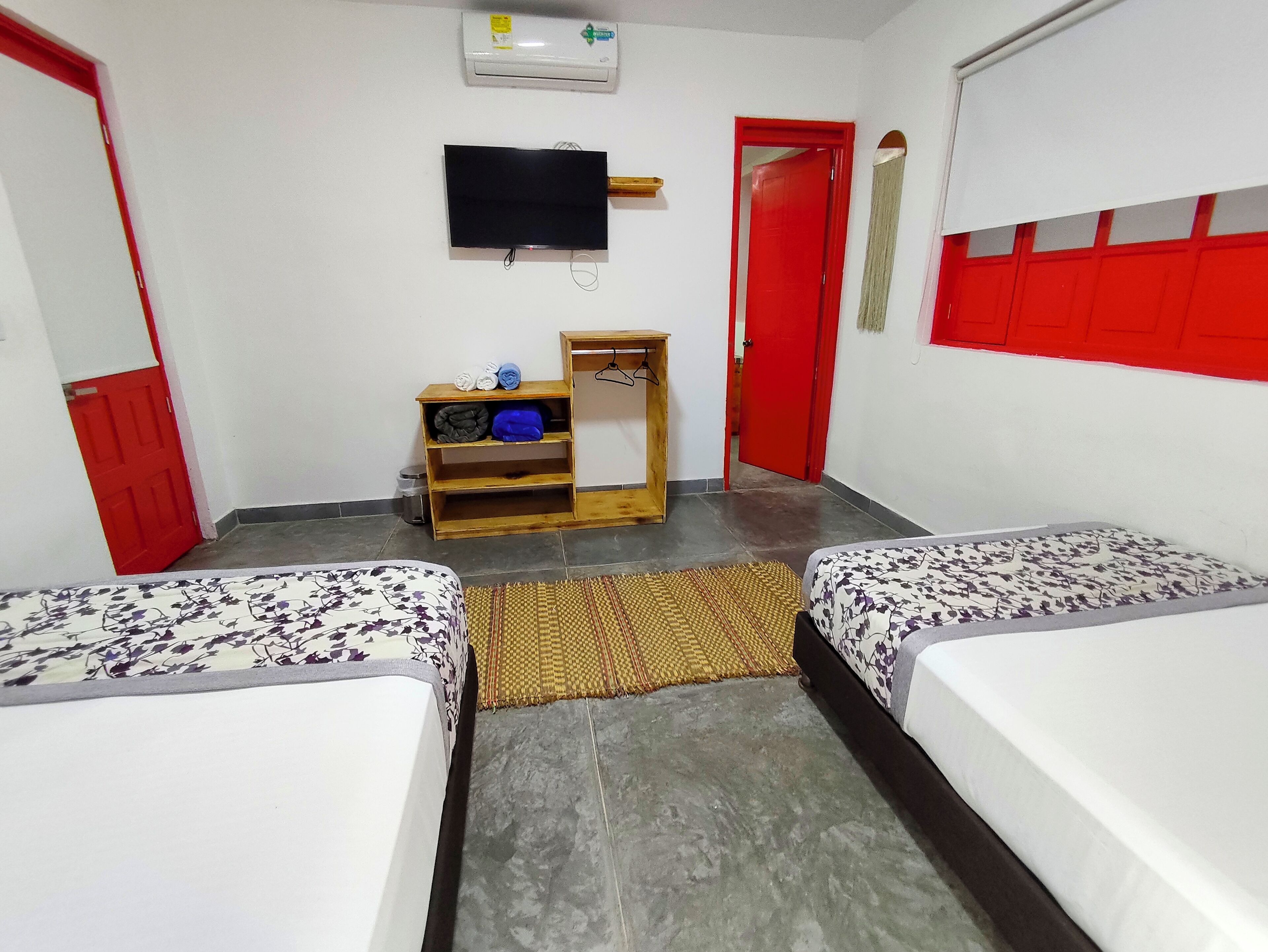 A room with two beds, a TV, a shelf with clothes, and a red door.