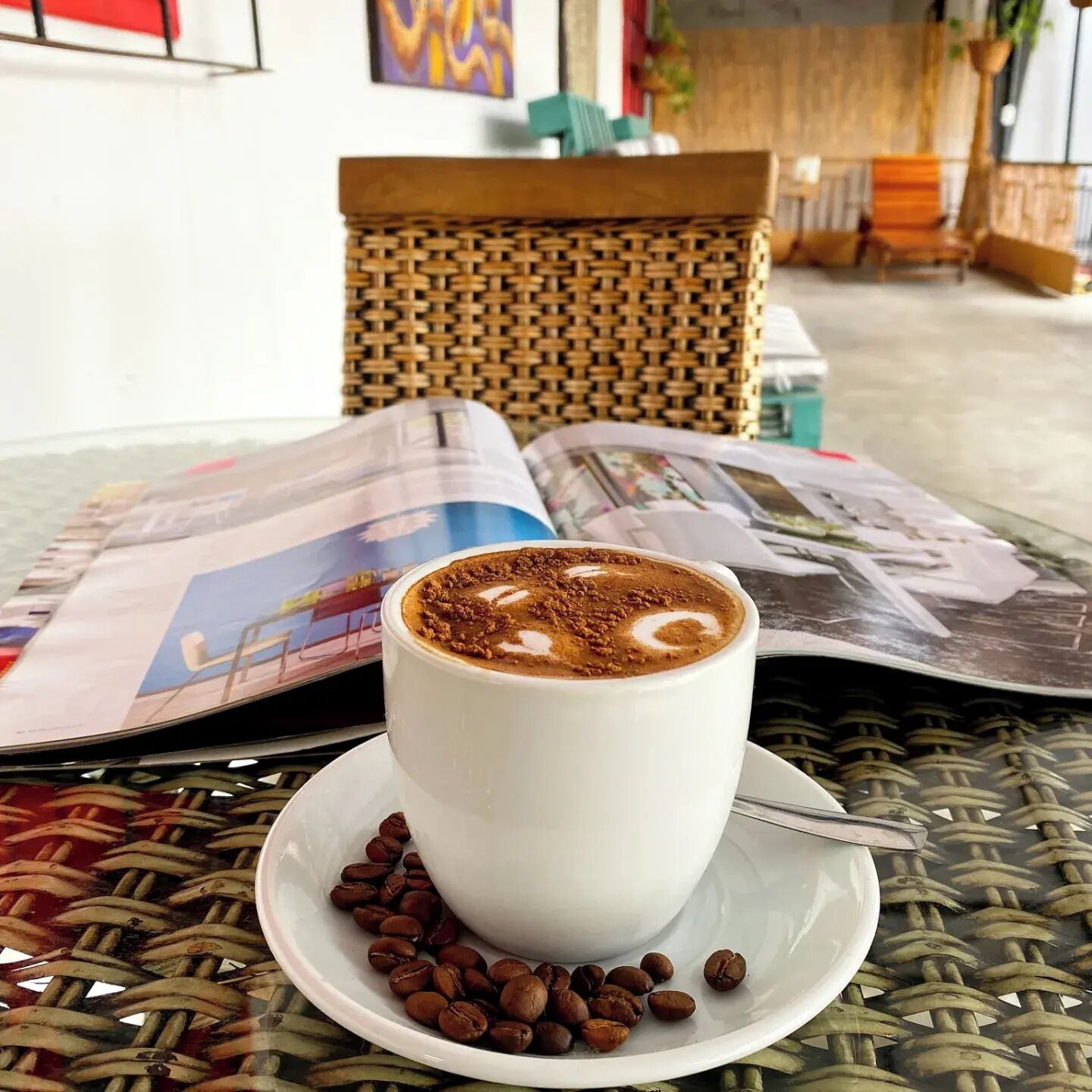 A white cup of coffee with foam on a white saucer, placed on a woven table with a newspaper underneath.
